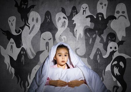 Excited girl hiding under blanket from imaginary ghosts. Scared kid sitting in his bed at home. Little girl afraid of fantasy monsters. Covered child in pajamas not sleep at night. Halloween holiday.の写真素材