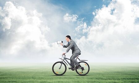 Businessman with megaphone in hand on bike at sunny day. Promotion and announcement. Corporate employee in business suit riding bicycle on green meadow. Handsome cyclist on background of blue sky.の写真素材