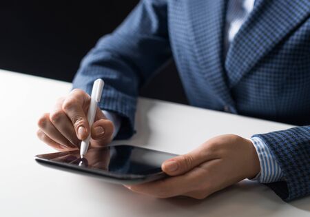 Man in business suit sitting at desk and working at tablet computer. Close-up of human hands holding pen and tablet gadget. Manager at workplace in office. Business and digital technology conceptの写真素材