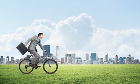 Young man riding bicycle on green grass. Businessman on bike hurry to somewhere. Corporate employee in grey business suit with suitcase on cycle outdoor. Male cyclist on green meadow at sunny dayの写真素材