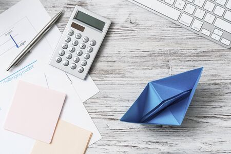 Modern office workspace with blue paper ship. Flat lay wooden desk with computer keyboard, calculator and documents. Creative marketing and advertising agency. Businessman leadership and directionの写真素材