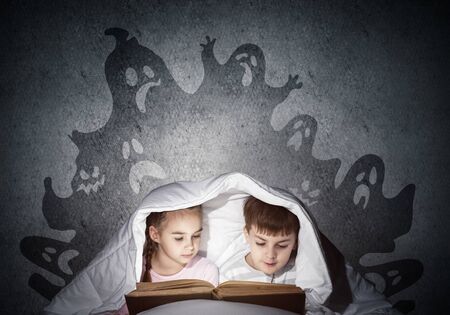 Engrossed little kids reading book in bed. Brother and sister together hiding under blanket. Fearful children in pajamas and imaginary monsters on background of grey wall. Child reading fairy tales.の写真素材
