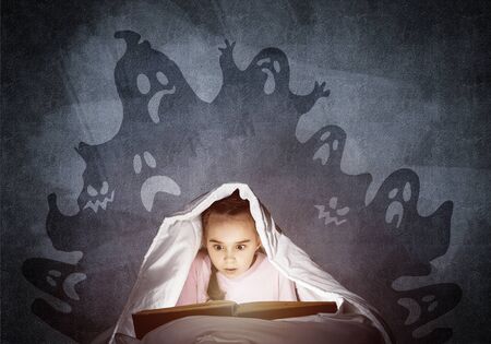 Scared little girl reading fairytales in bed. Child with flashlight hiding under blanket. Kid can not sleep at night. Girl in pajamas and frightening ghosts back on wall. Halloween holiday conceptの写真素材