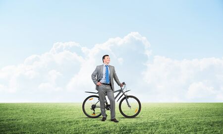 Smiling businessman with bicycle on background of blue cloudy sky. Young man in grey business suit and tie with bike relaxing outdoors. Happy handsome cyclist on green field. Healthy active lifestyleの写真素材