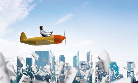 Businessman in aviator hat and goggles driving propeller plane above downtown. Papers falling down on background of skyscrapers. Funny man having fun in small airplane. Blue sky with clouds.の写真素材
