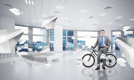 Smiling man in business suit standing with bike at comfortable conference room. Happy businessman with bicycle at modern office interior with big windows. Loft coworking space with flying paper planesの写真素材