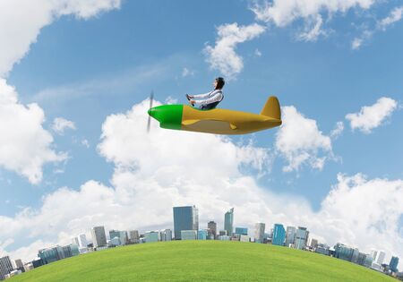 Happy businessman in leather helmet flying in propeller plane. Aviator driving small airplane above business center. Rounded city skyline with green grass, blue sky and modern office buildings.の写真素材