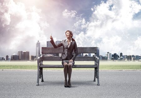 Business woman taking selfie photo or chatting with smartphone. Attractive girl using mobile phone on wooden bench. Mobile marketing and communication. Modern cityline panorama in sunny dayの写真素材