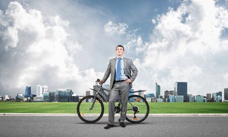 Young man wearing business suit and tie standing on asphalt road with bike. Businessman with bicycle on background of business center. Outdoors male cyclist holding bicycle, having break in riding.の写真素材