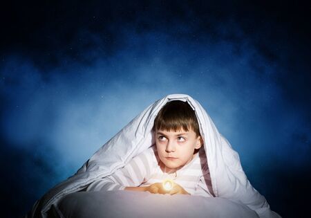 Frightened child with flashlight hiding under blanket. Scared kid lying in his bed at home. Fear of the dark. Little boy can not sleep at night. Portrait of boy in pajamas on background of starry skyの写真素材