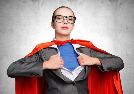 Attractive young business lady in red hero cape on grey wall background. Portrait of business woman super heroine. Career development and leadership. Confident super woman ready for new challenges.の写真素材
