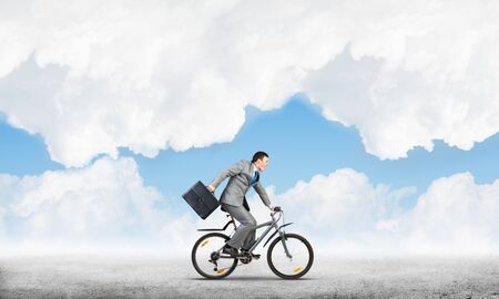 Man wearing business suit riding bicycle outdoor. Cyclist with suitcase on background of blue sky. Time management and business activity. Businessman hurrying to work. Business competition and careerの写真素材
