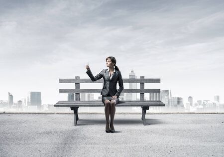 Business woman taking selfie photo or chatting with smartphone. Attractive girl using mobile phone on wooden bench. Mobile marketing and communication. Modern cityline panorama with cloudy sky.の写真素材