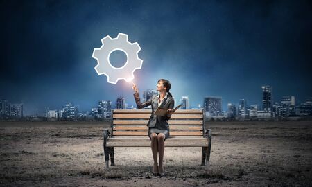 Young woman holding open book on wooden bench outdoor. Beautiful girl finger pointing at gearwheel symbol in night sky. Modern cityscape panorama at night. Engineering and development conceptの写真素材
