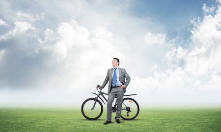 Smiling businessman with bicycle on background of blue cloudy sky. Young man in grey business suit and tie with bike relaxing outdoors. Happy handsome cyclist on green field. Healthy active lifestyleの写真素材