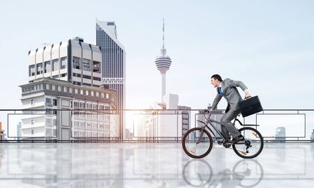 Man wearing business suit riding bicycle on penthouse balcony. Young cyclist with suitcase on background of business district. Terrace with modern downtown view. Time management and business activityの写真素材