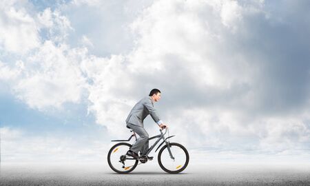 Businessman commuting to work by bike. Man in business suit riding bicycle on road. Handsome cyclist on background of blue sky. Nature landscape with copy space. Eco-friendly transport and active lifeの写真素材