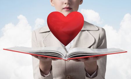 Beautiful woman showing red heart above opened notebook. Love and tenderness, valentines holiday and fall in love concept. Elegant girl in white business suit with open book on background of skyscape.の写真素材