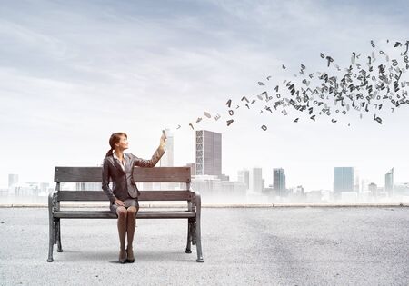 Young woman with smartphone sending text message. Girl sitting on wooden bench on background modern city panorama. Various letters flying out from mobile phone. Mobile marketing and communicationの写真素材