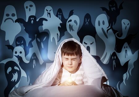 Young boy hiding under blanket from imaginary ghosts. Scared kid with closed eyes lying in his bed at home. Little boy afraid of fantasy monsters. Covered child in pajamas not sleep at night.の写真素材
