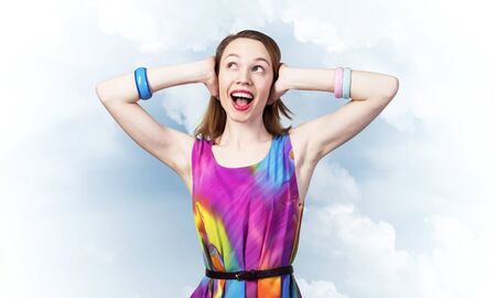 Redhead beautiful girl closing her ears with hands. Charming lady in bright colorful dress and bracelets against cloudy sky background. Portrait of happy young woman singing and smiling broadlyの写真素材