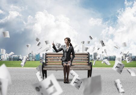 Business woman taking selfie photo or chatting with smartphone. Attractive girl using mobile phone on wooden bench. Mobile marketing and communication. Cityline panorama with falling paper documentsの写真素材