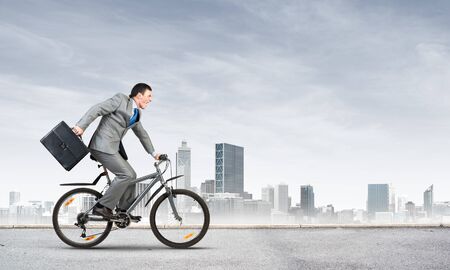 Young man riding bicycle on highway. Businessman on bike hurry to work. Corporate employee wearing business suit with suitcase on cycle outdoors. Cyclist on background of urban architecture.の写真素材