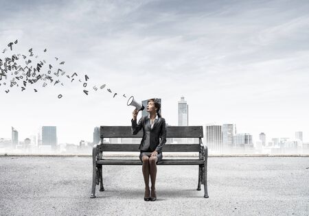 Young woman with megaphone sitting on wooden bench. Female speaker advertising new service or product on city panorama. Various letters flying out from loudspeaker. Business marketing and announcementの写真素材