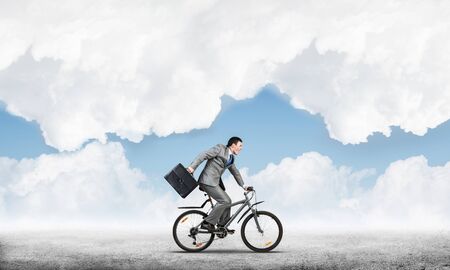 Man wearing business suit riding bicycle outdoor. Cyclist with suitcase on background of blue sky. Time management and business activity. Businessman hurrying to work. Business competition and careerの写真素材