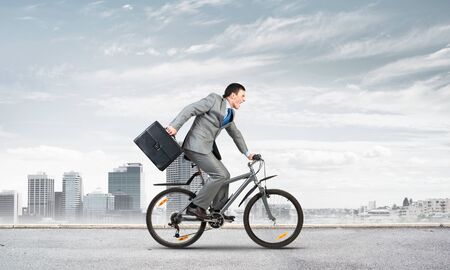 Young man riding bicycle on highway. Businessman on bike hurry to work. Corporate employee wearing business suit with suitcase on cycle outdoors. Cyclist on background of urban architecture.の写真素材