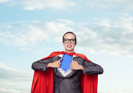 Portrait of business woman super heroine. Attractive young business lady in suit with red cloak on blue sky background. Successful and confident super woman in glasses ready for new challenges.の写真素材