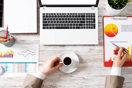 Top view modern office workspace with human hands. Flat lay white wooden desk with laptop keyboard, business infographics and cup of coffee. Businessman hands working at computer mockup.の写真素材