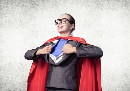 Portrait of business woman super heroine tearing off her shirt. Smiling business lady with closed eyes in red hero cape on grey wall background. Brave super woman dreaming about new winsの写真素材