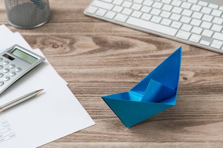 Modern office workspace with blue paper ship. Corporate strategy and successful solution. Flat lay wooden desk with computer keyboard, calculator and documents. Creative innovation and leadershipの写真素材