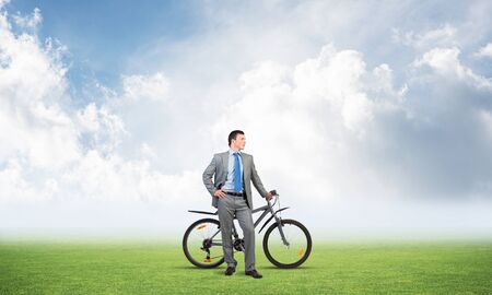 Young man in grey business suit and tie standing on green grass with bike. Cheerful businessman with bicycle on background of blue cloudy sky. Male cyclist looking confidently. Eco friendly transportの写真素材