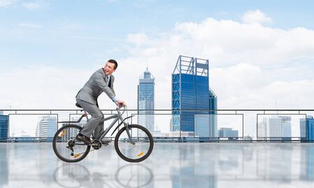 Man wearing business suit riding bicycle on penthouse balcony. Handsome cyclist looking back on background of urban architecture. Terrace with modern city center view. Outdoor fun and activity.の写真素材