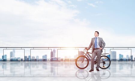 Young man wearing business suit standing on balcony with bike. Businessman with bicycle on background of blue sky above megalopolis. Male cyclist holding bicycle on terrace with modern downtown view.の写真素材