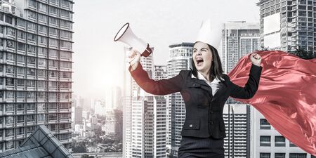 Young confident businesswoman wearing red cape against modern city background screaming in megaphoneの写真素材