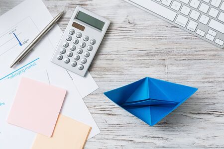 Modern office workspace with blue paper ship. Corporate strategy and smart solutions. Flat lay wooden desk with computer keyboard, calculator and documents. Creative marketing and advertising agencyの写真素材