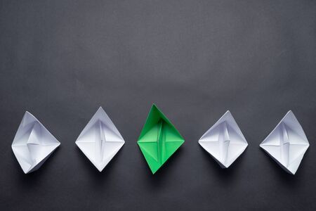 Row of paper ships on black background. Individual motivation and direction. Creative innovation and leadership. Flat lay green origami boat in group of white boats. Social marketing layout.の写真素材