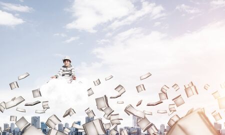 Young little boy keeping eyes closed and looking concentrated while meditating on cloud among flying books with cityscape view on background.の写真素材
