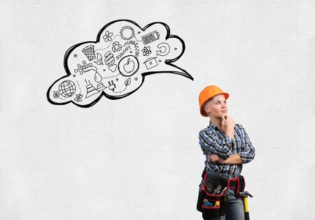 Concentrated female construction worker in safety helmet standing with folded arms. Pensive young technician with speech bubble above head on white background. Business idea generation conceptの写真素材