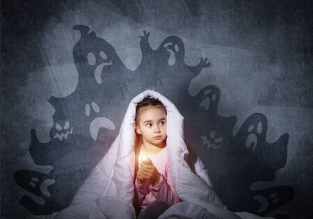 Scared girl with flashlight hiding under blanket. Frightened kid sitting in his bed at home. Night terrors of child. Beautiful girl in pajamas and boo ghosts silhouettes back on wall.の写真素材