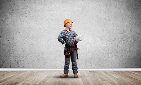 Attractive female architect in hardhat standing with technical blueprints. Portrait of young worker in checkered blue shirt and jeans standing in empty room. Interior design and renovation service.の写真素材