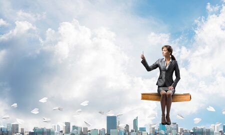 Attractive girl in business suit sitting on big book and flying in blue sky. Young woman taking selfie photo with smartphone. Pretty businesswoman on background cityscape panorama with paper planes.の写真素材