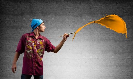 Young artist painting orange brush stroke. Male painter in dirty shirt and bandana with paintbrush on grey wall background. Creative hobby and artistic occupation. Art classes concept with copy space.の写真素材