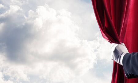Human hand in glove opens red velvet curtain on blue sky backgroundの写真素材