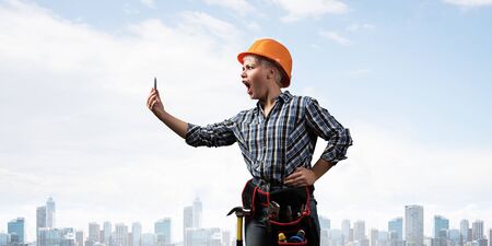 Expressive blonde woman in workwear and hardhat shouting into smartphone. Portrait of young emotional worker in checkered blue shirt standing with open mouth. Business communication and conversation.の写真素材
