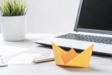 Businessman workspace with office wooden desk and yellow paper ship. Flat lay table with plant in pot and laptop computer. Close up orange origami boat. Creative and innovative solution for business.の写真素材