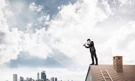 Young determined businessman standing on house roof and looking in spyglass. Mixed mediaの写真素材